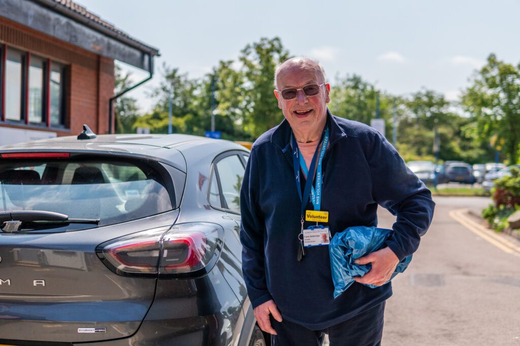 A man stnading outside his car, with a volunteering badge